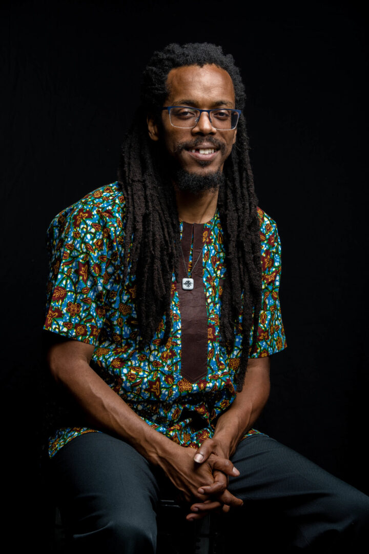 Photo of Bomani Armah dressed in a multii colored shirt sitting down with a black background