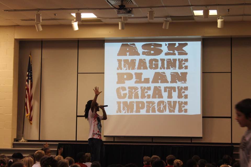 A photo of Bomani at a screen with the words: ask, imagine, plan, create, improve