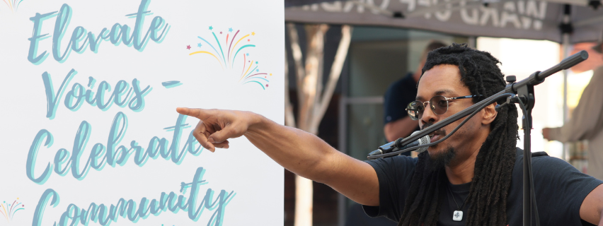 Bomani Armah at Fresh Farms in Silver Spring, he is at the micorphone and there is a sign behind his pointing finger saying Elevate Voices, Celebrate Community